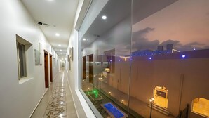 Corredor longo e bem iluminado, com paredes de vidro, que dá para uma varanda com vista para a paisagem urbana ao entardecer.