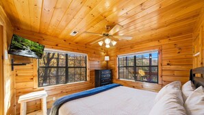 Ein Schlafzimmer einer Holzhütte mit einem Bett, zwei Fenstern, einem Deckenventilator und einer Kommode.