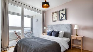 A modern bedroom with a large bed, two bedside tables with lamps, a balcony with a chair, and abstract wall art.