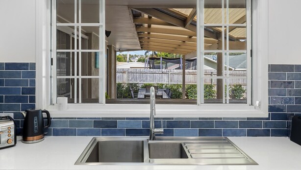 A kitchen with a double sink, a window with a view of a backyard, and blue-tiled walls. - Beach House With Pool, Pets Welcome! (Coolum Beach)