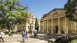 Una plaza pública con una fuente, un gran edificio de estilo cl ásico con columnas, y gente caminando.