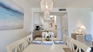 A dining area with a chandelier, white chairs, a table with books and a vase of flowers, and a kitchen in the background. - Uncrowded Corner Beachfront 3BR w/3 New Spa Baths (Gulf Shores)
