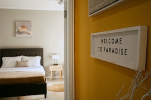 Un dormitori amb un llit, una tauleta de nit i un cartell al mur que diu 'WELCOME TO PARADISE'.