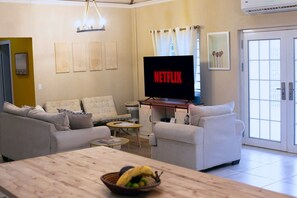 A living room with a television displaying 'NETFLIX,' a sofa, a coffee table, and a dining table with a fruit bowl. - The Nest Villa - Sleeps 16, 5BR Family Pool, Swings, BBQ, Gated Estate (Canaan, Tobago)