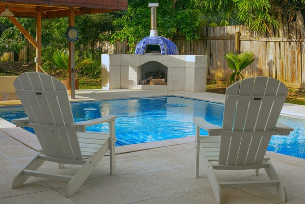 Una piscina con dos sillas Adirondack, un pozo de fuego de piedra y una valla de madera al fondo.