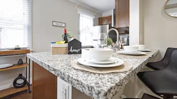Una cocina con encimeras de granito, armarios de madera y una silla negra. Hay platos y tazones blancos sobre la encimera.