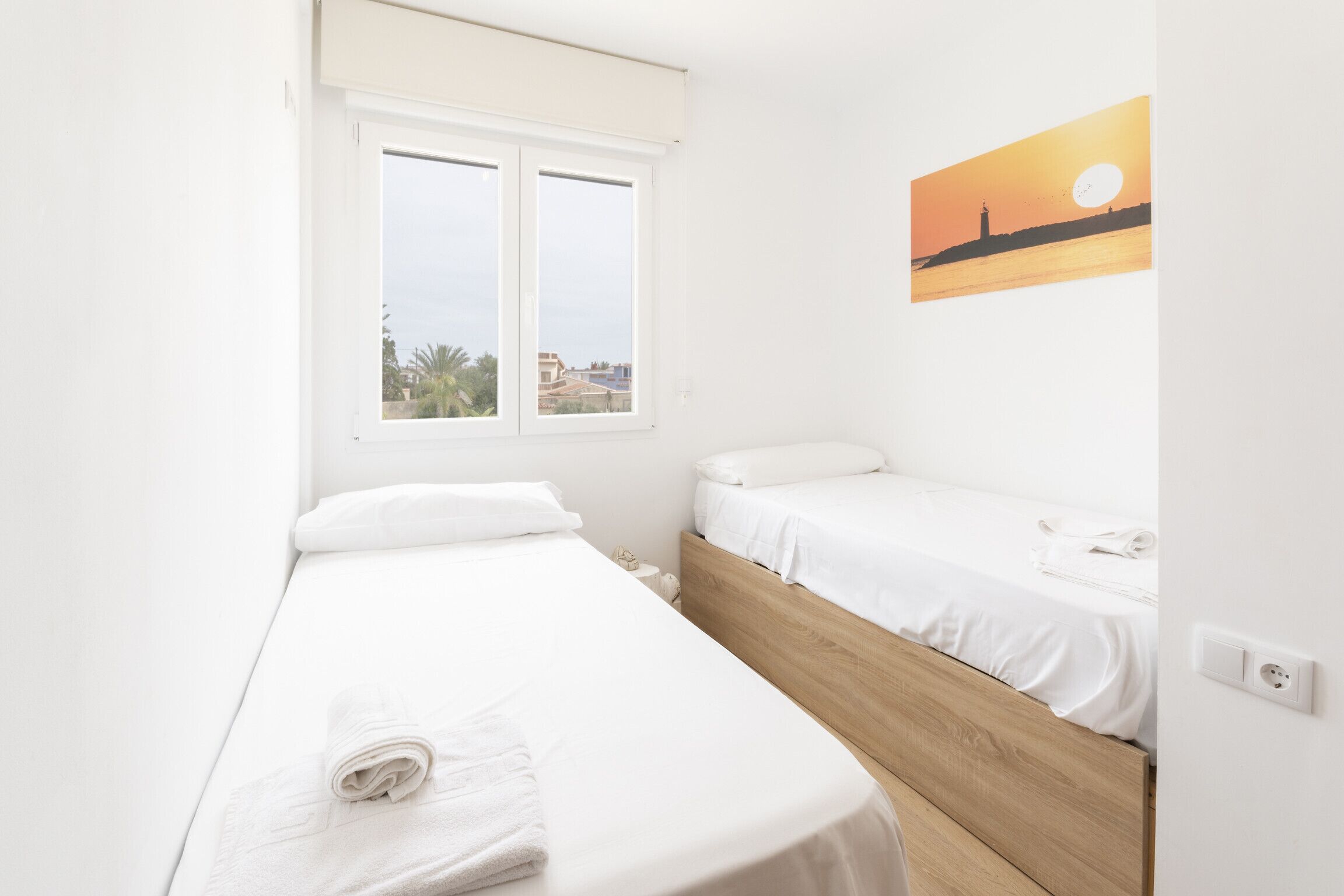 A bedroom with two single beds, a window with a view of buildings, and a sunset painting on the wall.