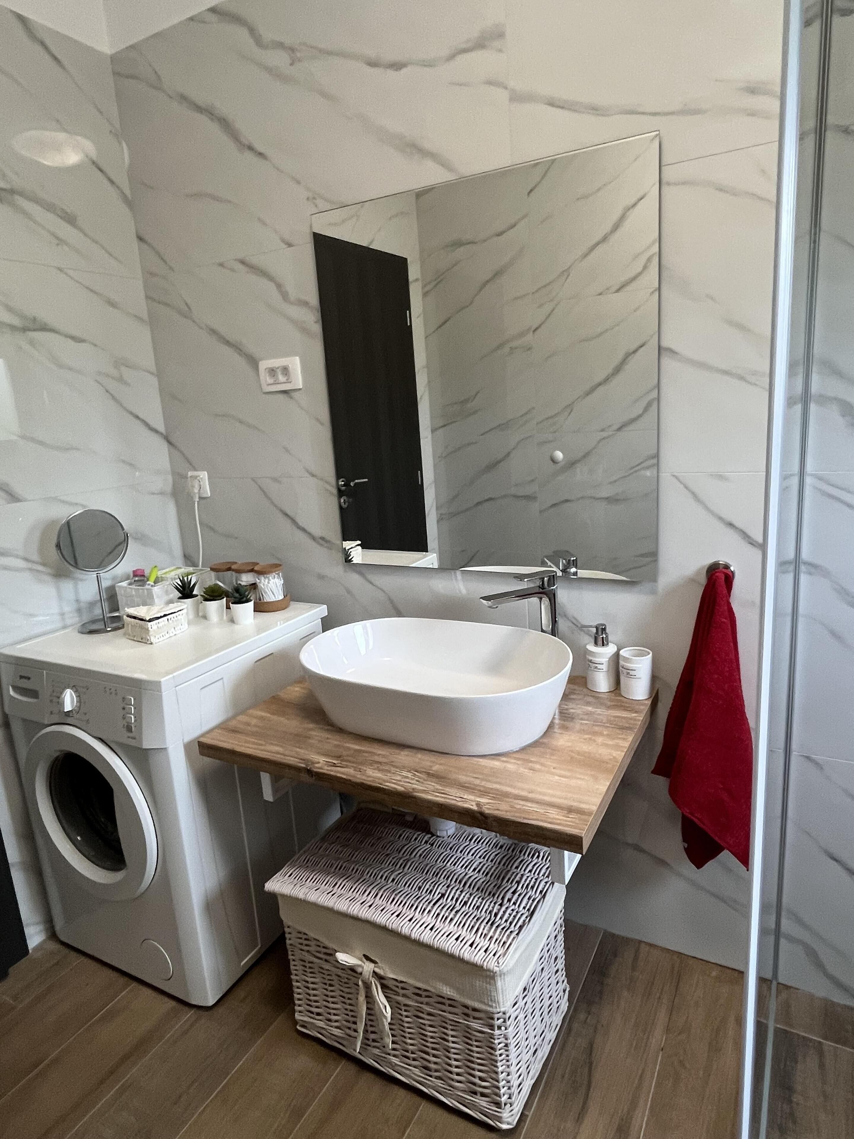 A modern bathroom with a marble wall, a large mirror, a white sink, a washing machine, and a red towel.