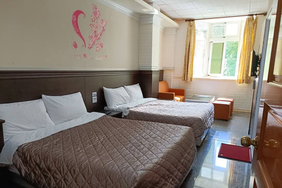 Quarto de hotel com duas camas, cabeceira de madeira, uma mesinha e uma janela com cortinas.