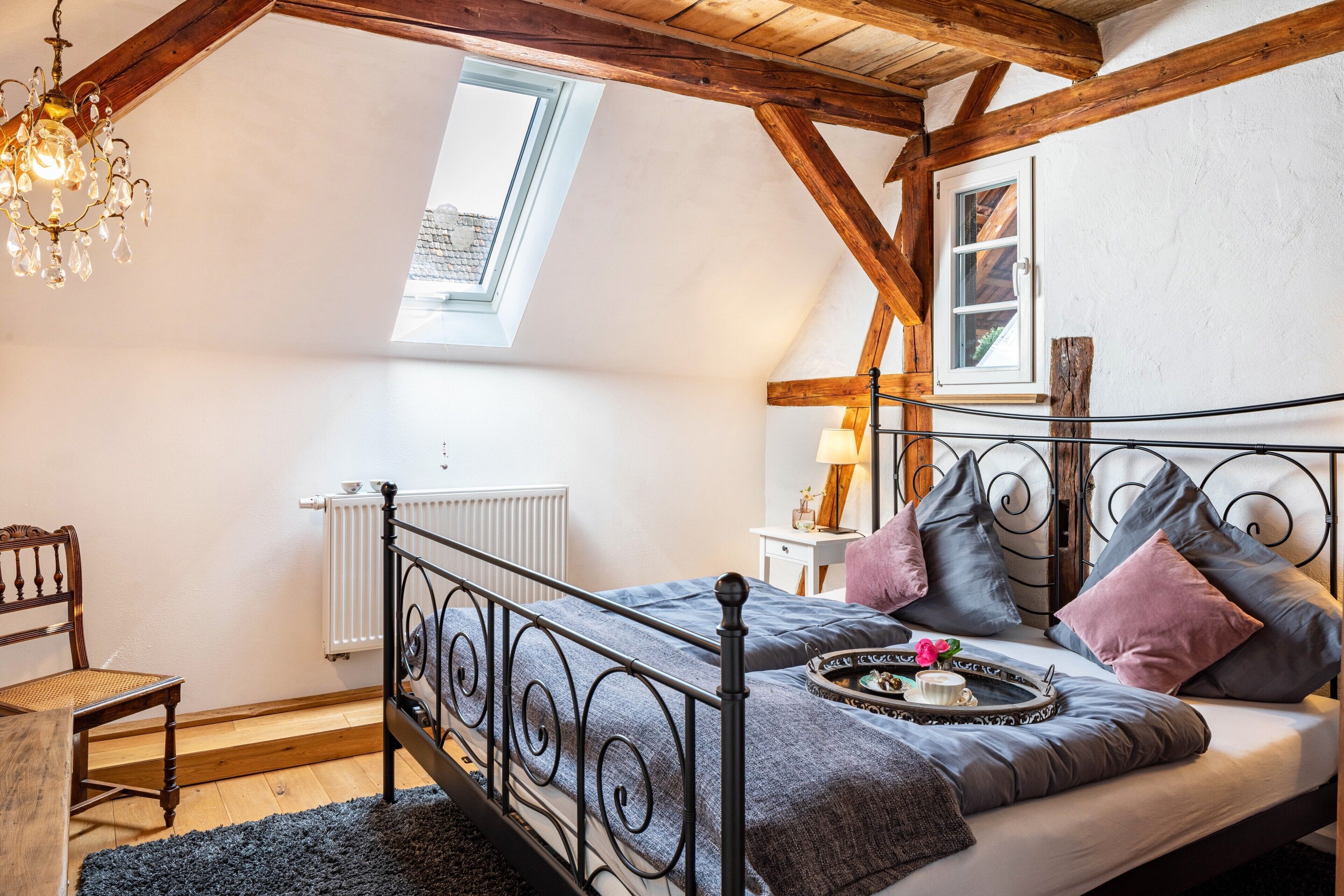 Una camera da letto con soffitto spiovente, un letto con struttura in metallo, una sedia in legno, un piccolo tavolo con una lampada e una finestra vista esterna.