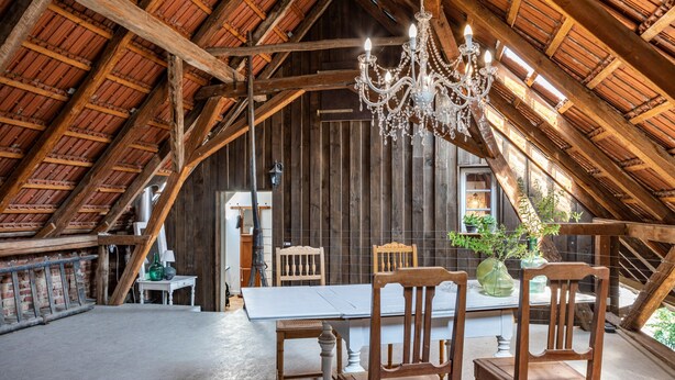 Een zolderkamer met een houten plafond, een kroonluchter, een eettafel en houten stoelen.