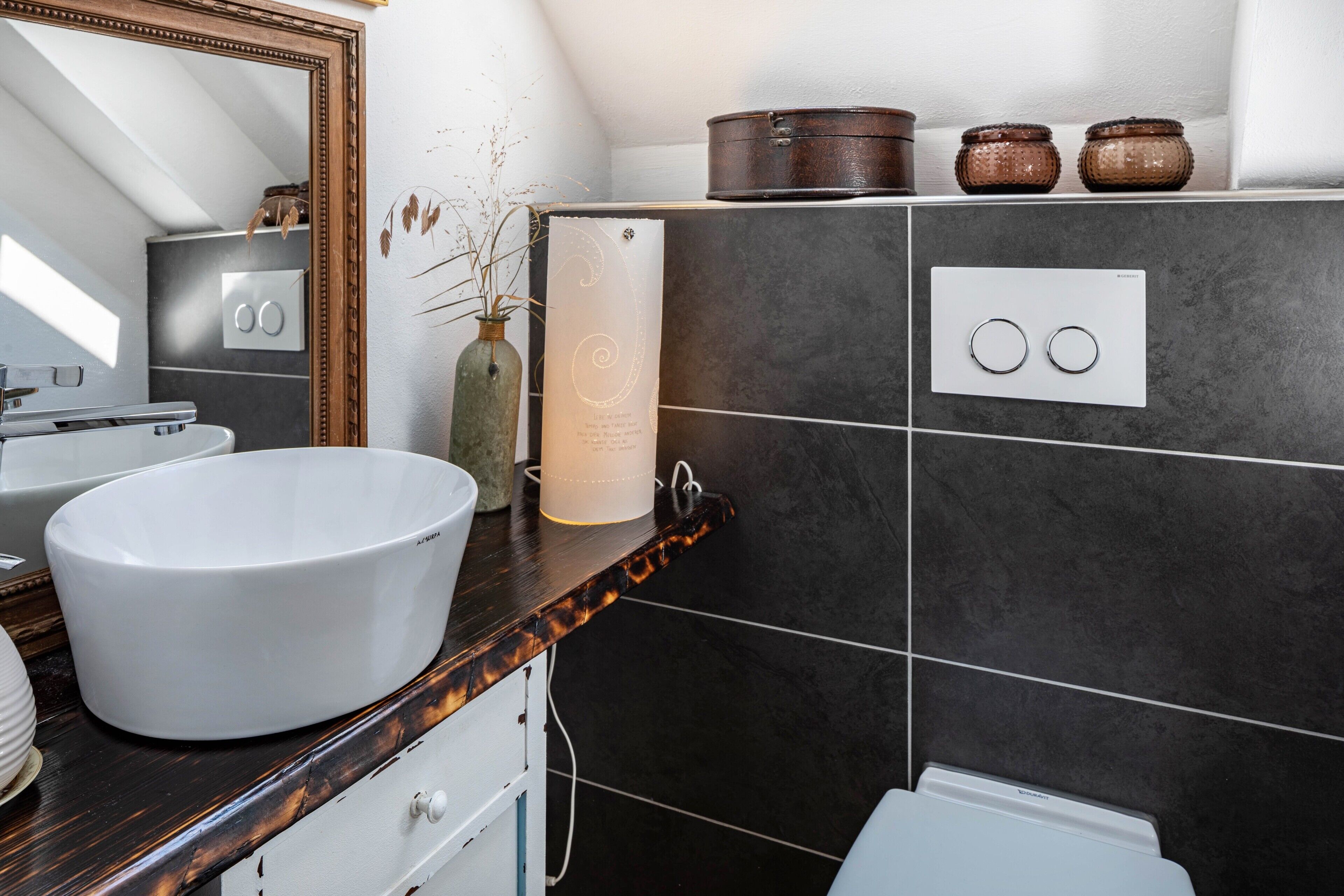 A modern bathroom with a white sink, dark tiled walls, a wooden countertop, and a mirror.