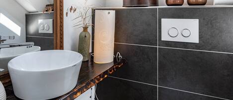 A modern bathroom with a white sink, dark tiled walls, a wooden countertop, and a mirror.