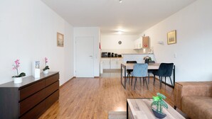 A modern living room with a wooden floor, a brown sofa, a dining table with chairs, a kitchen in the background, and a small potted plant on the coffee table.
