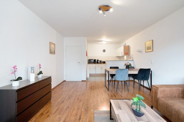 A modern living room with a wooden floor, a brown sofa, a dining table with chairs, a kitchen in the background, and a small potted plant on the coffee table.