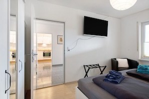 A bedroom with a bed, a TV mounted on the wall, a chair, and a view into the kitchen.
