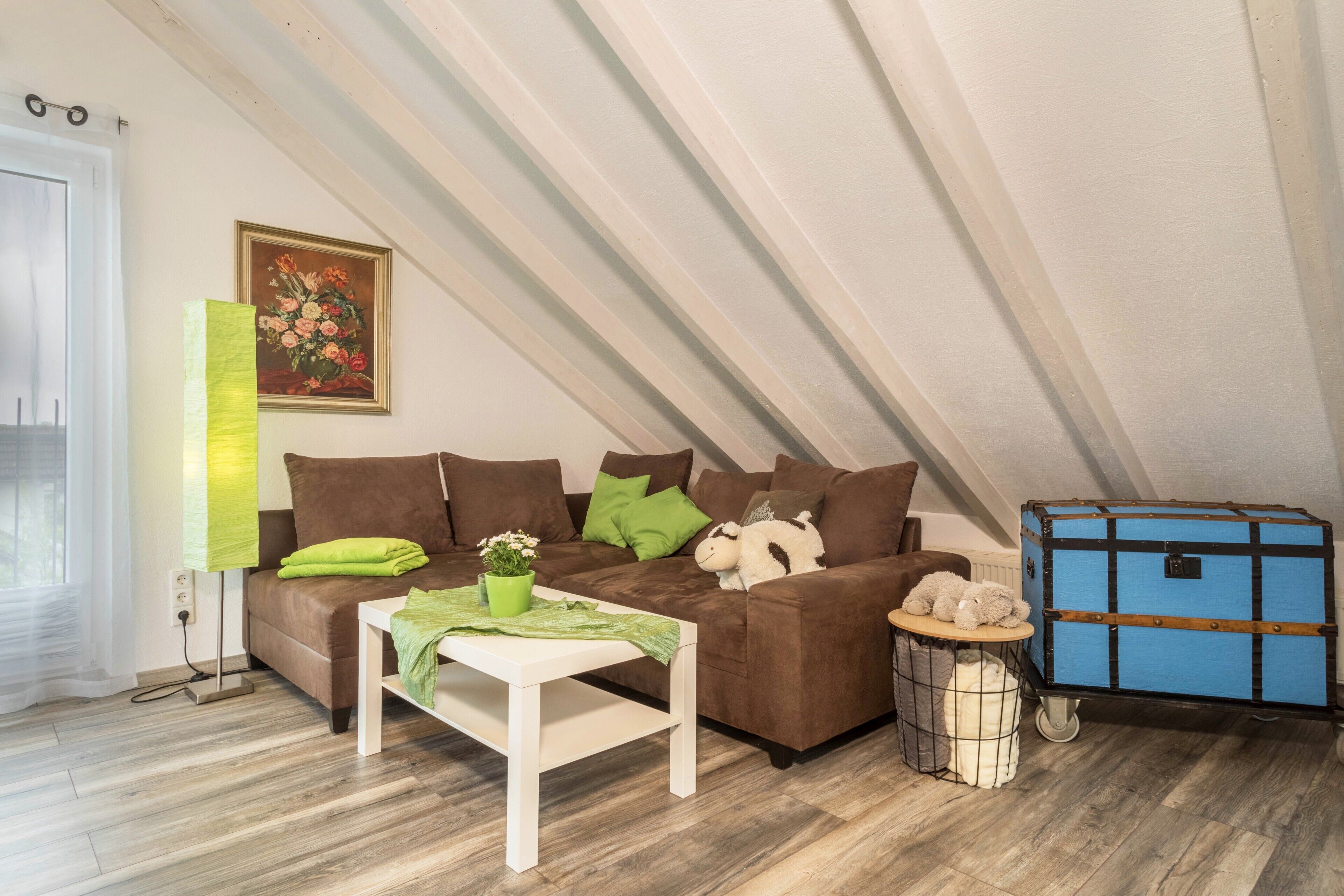 Une chambre mansardée chaleureuse avec un canapé marron, une petite table surmontée d’un coussin vert, une lampe, un tableau et une valise bleue.