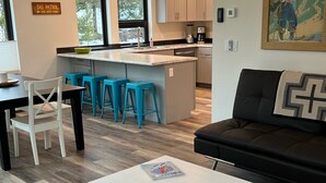 Een moderne woonkamer met een witte salontafel, een zwarte bank en een tapijt met een patroon. In de achtergrond is een keuken zichtbaar met een groot eiland en blauwe barkrukken.