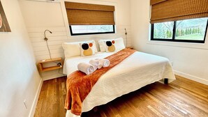 Un dormitorio con una cama grande, dos ventanas con persianas, piso de madera y una mesita de noche pequeña.