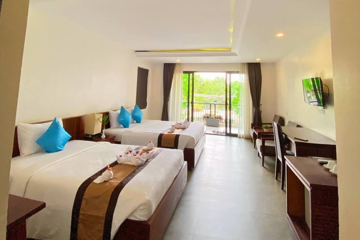A hotel room with two beds, a desk, and a view of greenery outside.