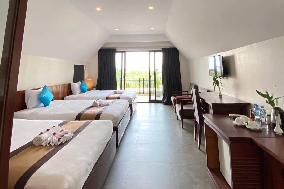 A hotel room with three beds, a desk, a TV, and a balcony with curtains.