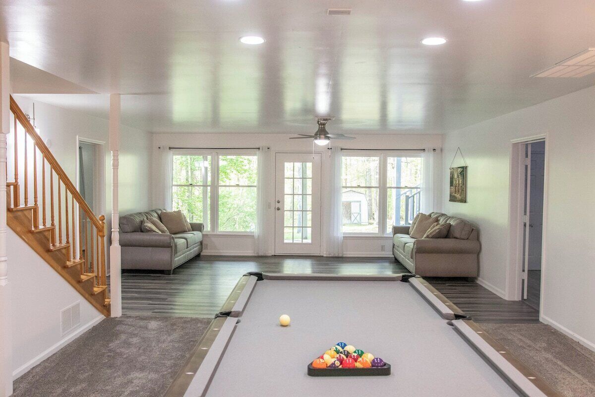 Amplia habitación con mesa de billar, dos sofás, una escalera y ventanas grandes.