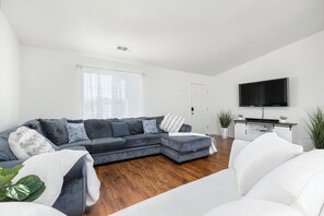 A modern living room with a grey sectional sofa, white armchairs, a flat-screen TV, and a wooden floor. - Bright & Comfy House Near NAS with Free Parking (Armona)