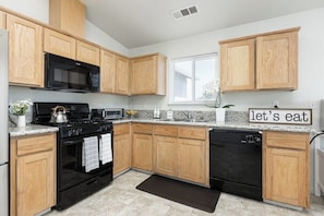 A modern kitchen with wooden cabinets, a black dishwasher, and a sign that says 'let's eat'. - Bright & Comfy House Near NAS with Free Parking (Armona)