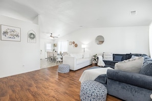 A modern living room with hardwood flooring, a white sofa, a blue sofa, and patterned ottomans. - Bright & Comfy House Near NAS with Free Parking (Armona)
