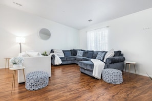 A modern living room with a sectional sofa, ottomans, a round mirror, and a small side table. - Bright & Comfy House Near NAS with Free Parking (Armona)