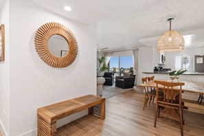 A modern living room with a wooden bench, a round mirror, and a dining area with wooden chairs and a table.