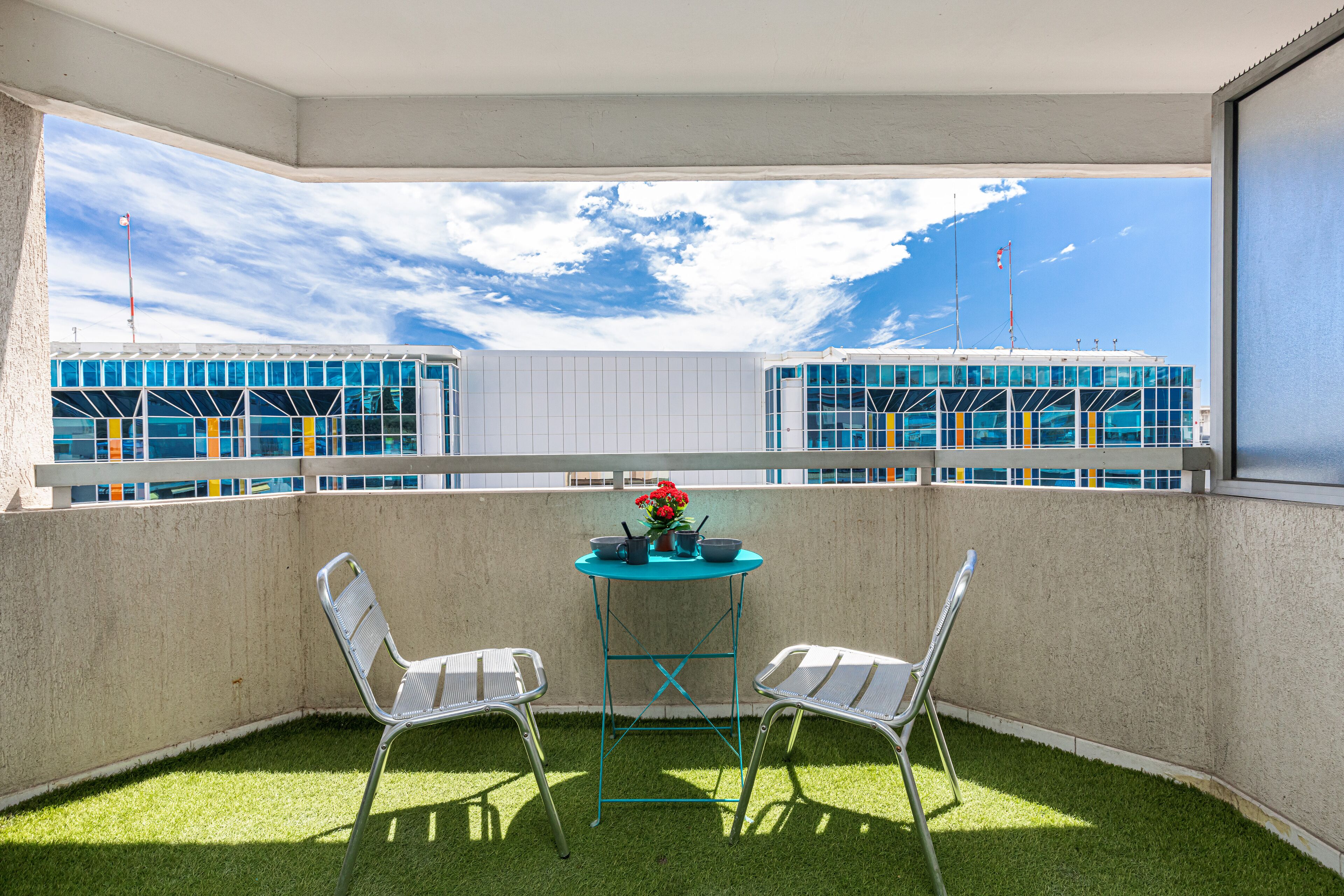 A balcony with two chairs, a small table with a flower arrangement, and a view of buildings in the background.