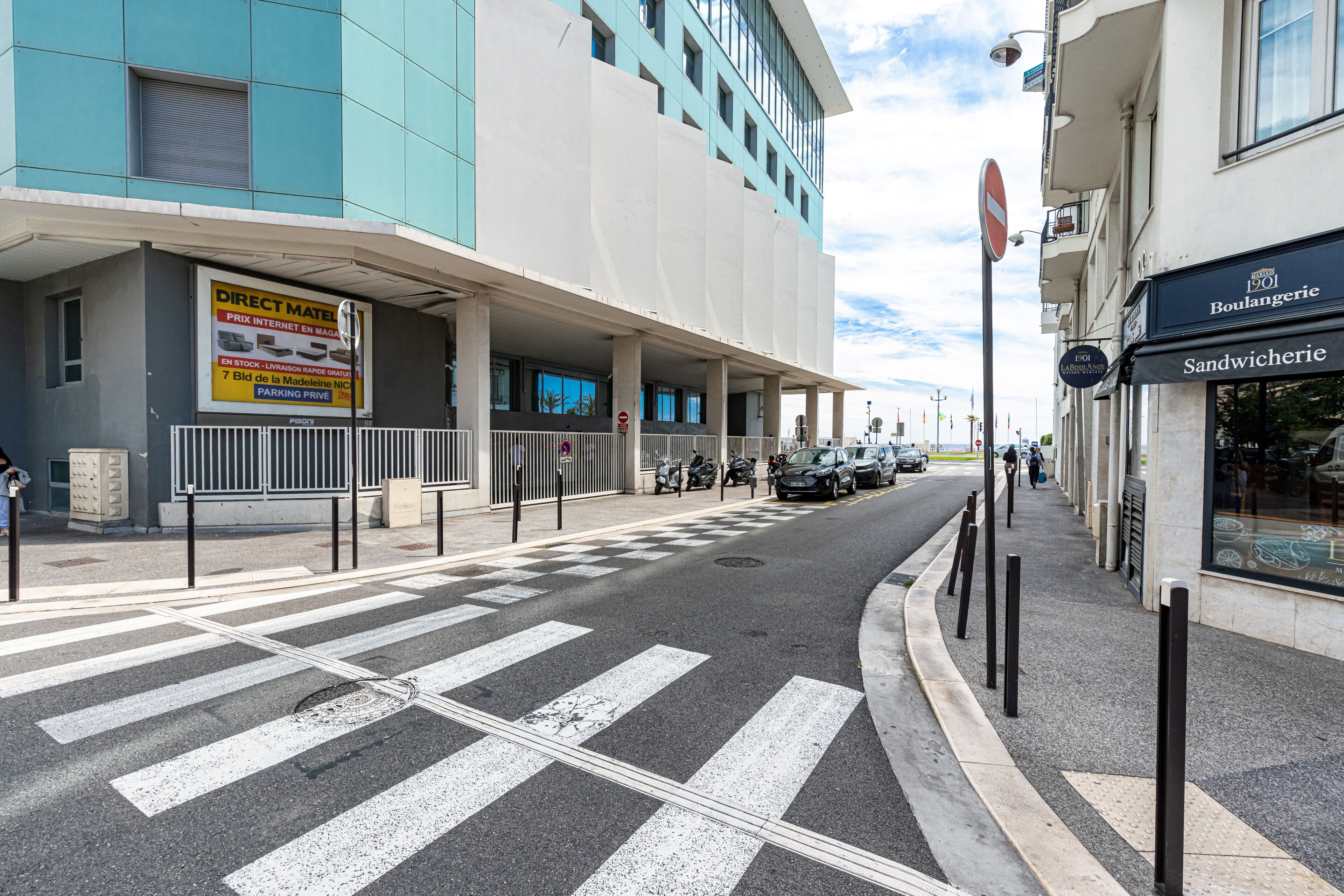 Une vue de rue avec un bâtiment portant l’inscription « Direct Matel » et un magasin appelé « Sandwicherie ».
