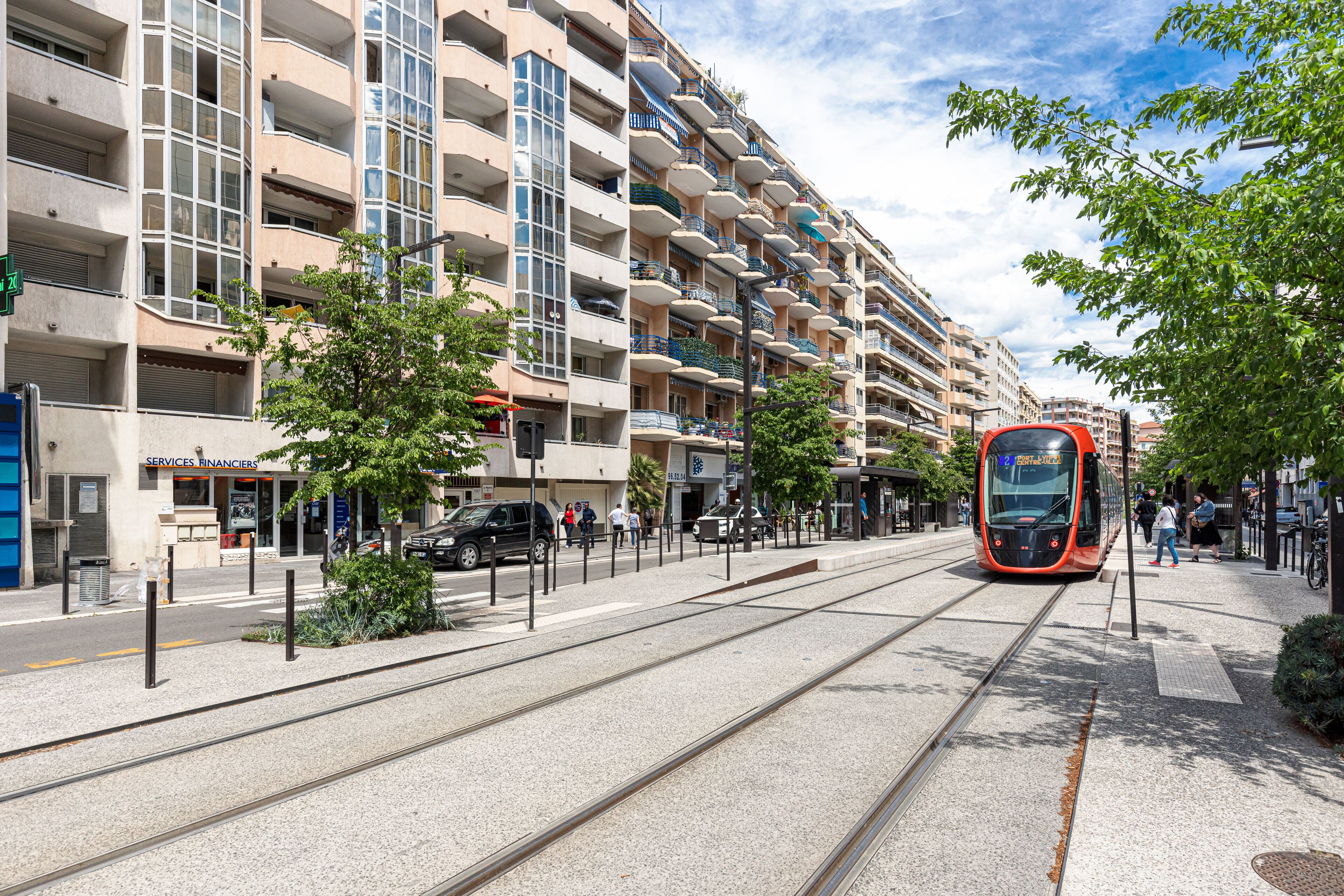 Een tram rijdt op rails, met op de achtergrond een meerlaags gebouw met balkons en een voetpad met voetgangers.