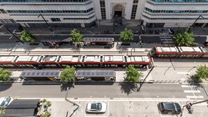 En bygning med flere etager, en midterindgang, en sporvognslinje og et parkeringsområde med biler og en bus.