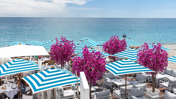 En restaurant ved stranden med udendørs siddepladser, blå og hvide striber parasoller samt pink buganvillea træer.