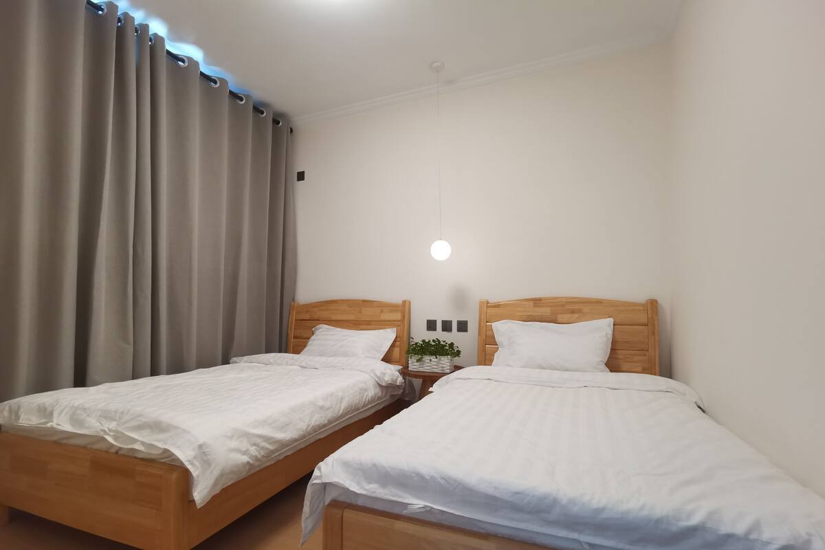 Une chambre avec deux lits en bois, des rideaux beiges, une petite table de chevet avec une plante et une lampe fixée au mur.