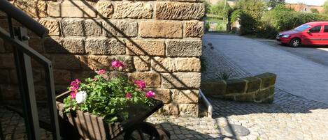 A stone wall with a wooden cart filled with flowers, a red car parked in the background, and a cobblestone path.