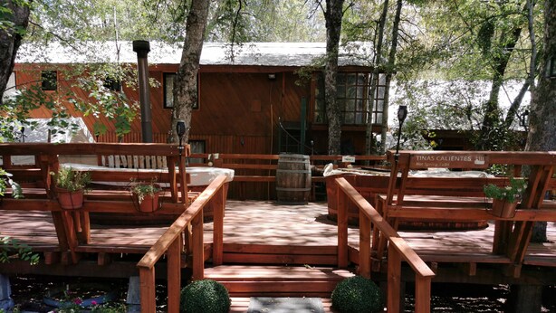 A wooden deck with benches and potted plants, a barrel, and a sign reading "Tinas Calientes Hot Spring Tubs".