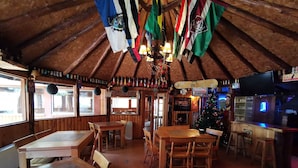 A rustic wooden interior with multiple tables and chairs, a Christmas tree, and various flags hanging from the ceiling.