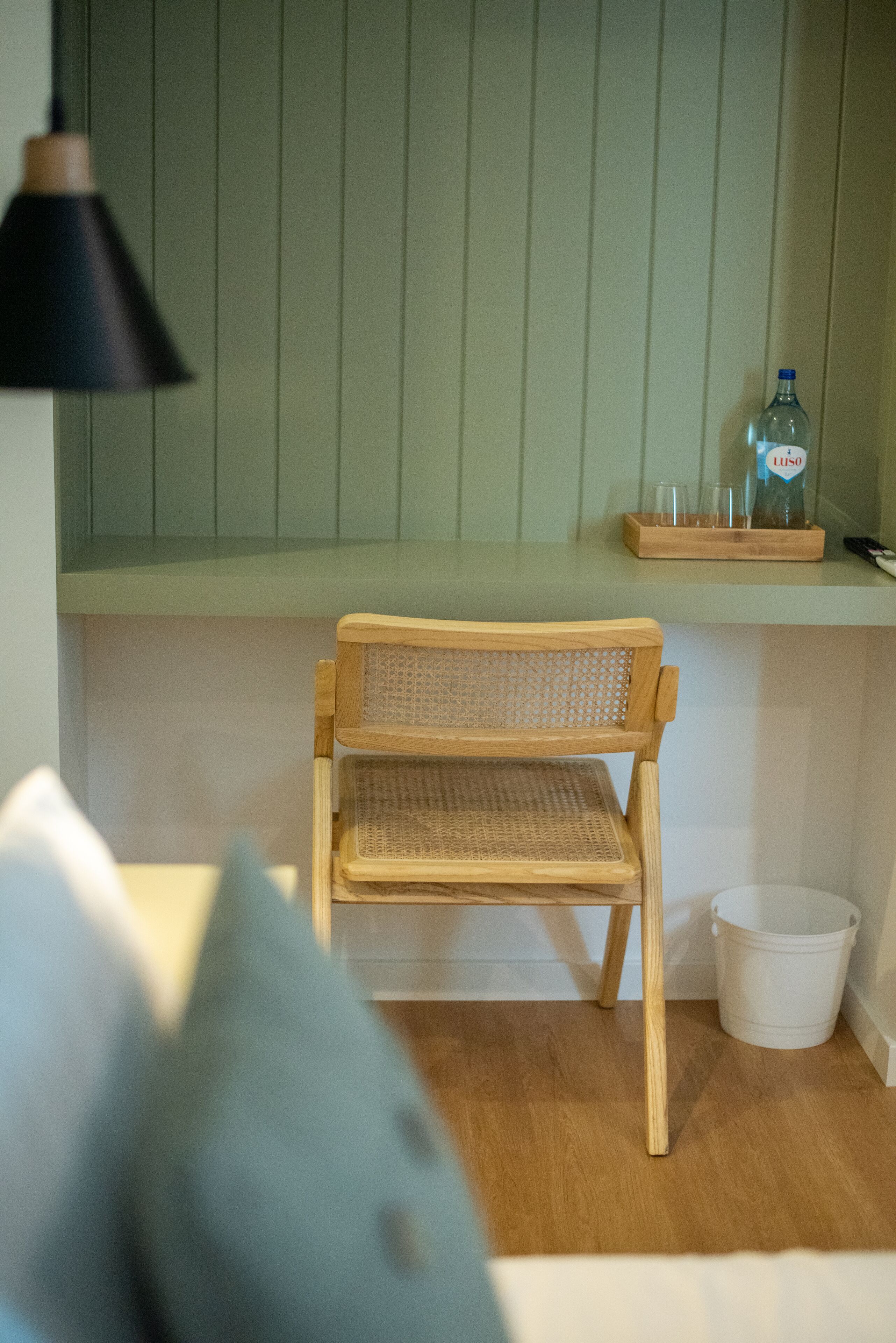A small wooden chair with a woven backrest and seat, positioned in front of a green wall with vertical paneling. A black pendant light hangs above, and a white bucket is visible on the floor to the right.