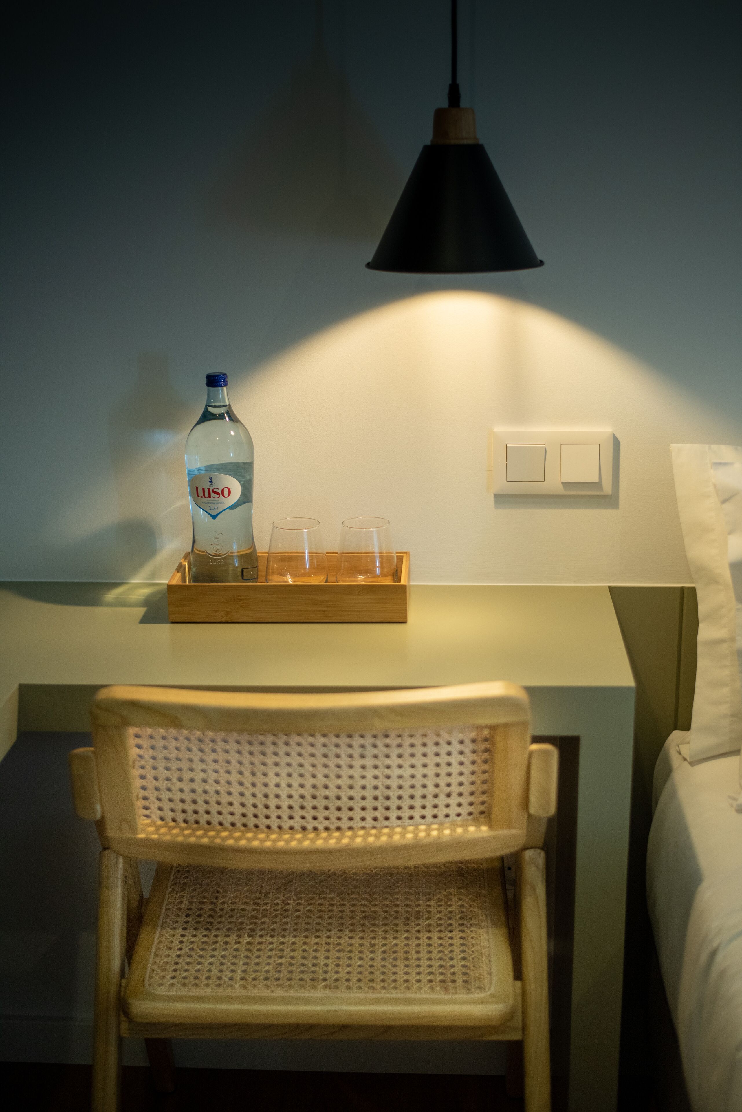 A hotel room with a wooden chair, a bedside table with a bottle of Luso and two glasses, a black pendant lamp, and a bed with white bedding.