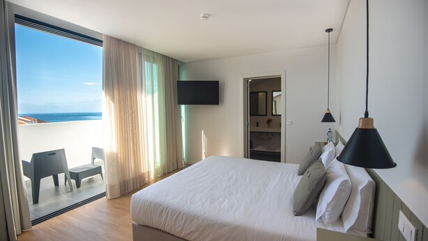 A bedroom with a large bed, a TV mounted on the wall, a balcony with a view of the sea, and a chair. - Olive Boutique Guesthouse  (Vila Franca do Campo)