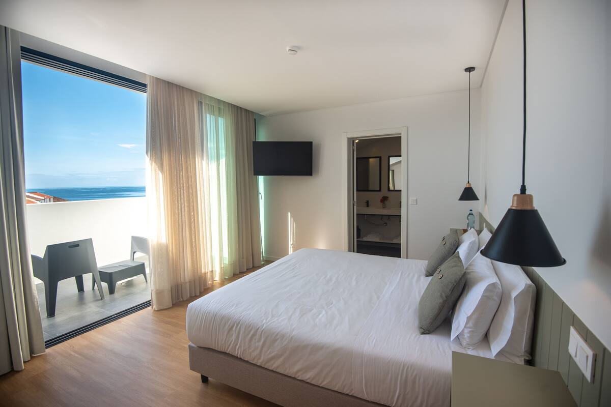 Ein Schlafzimmer mit einem großen Bett, einem an der Wand montierten Fernseher, einem Balkon mit Meerblick und einem Sessel.