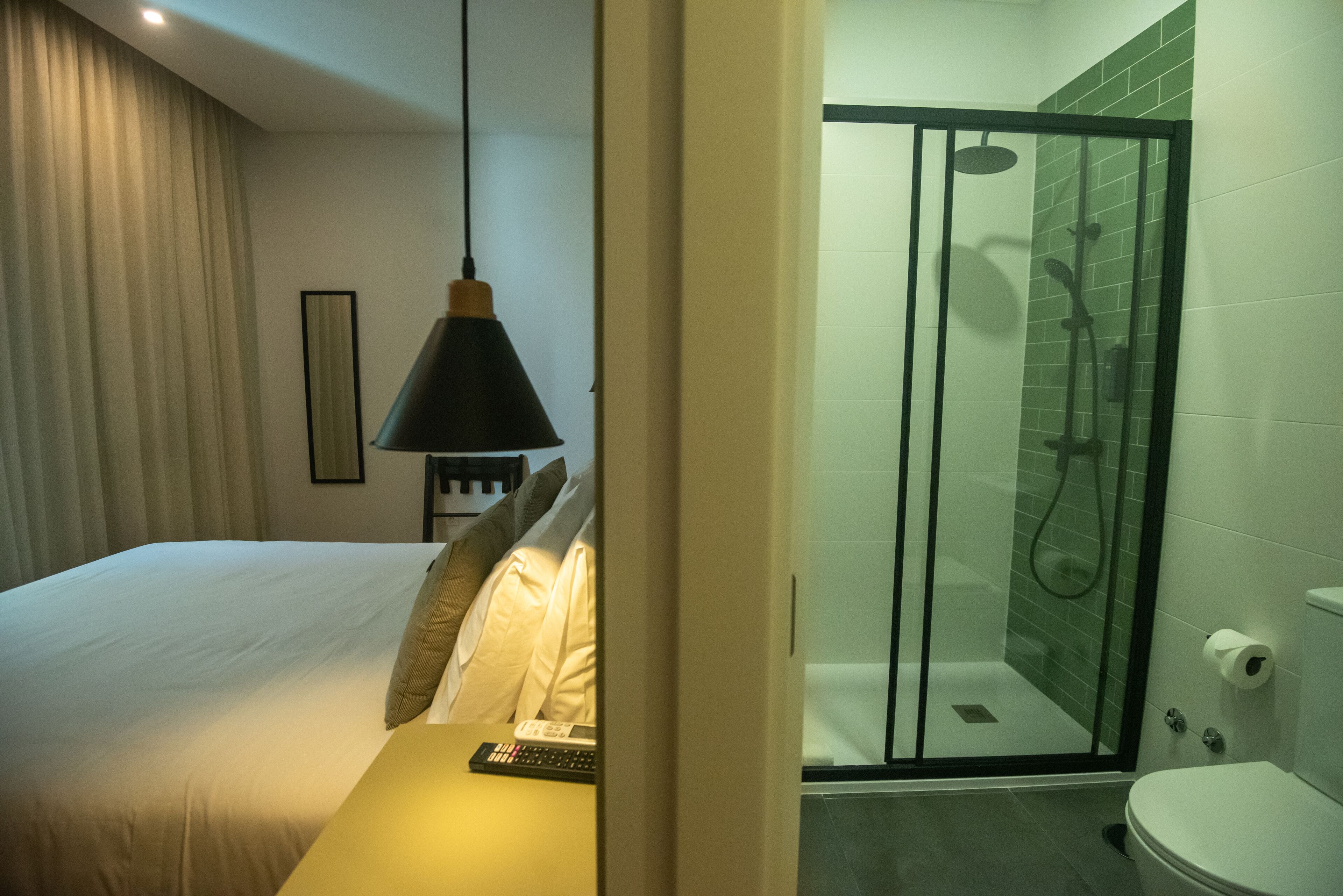 A hotel room with a bed, a bedside table with a remote control, a hanging lamp, a shower with a glass door, and a toilet.