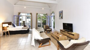 Ein geräumiges Wohnzimmer mit einem großen Bett, einem Sofa, einem Couchtisch und einem Fernseher. Große Fenster mit Vorhängen und ein Balkon, der durch die Fenster sichtbar ist.