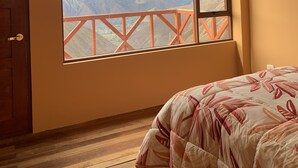Una habitación con un ventanal amplio que ofrece vistas a la montaña, una puerta de madera y una cama con colcha de motivos florales.