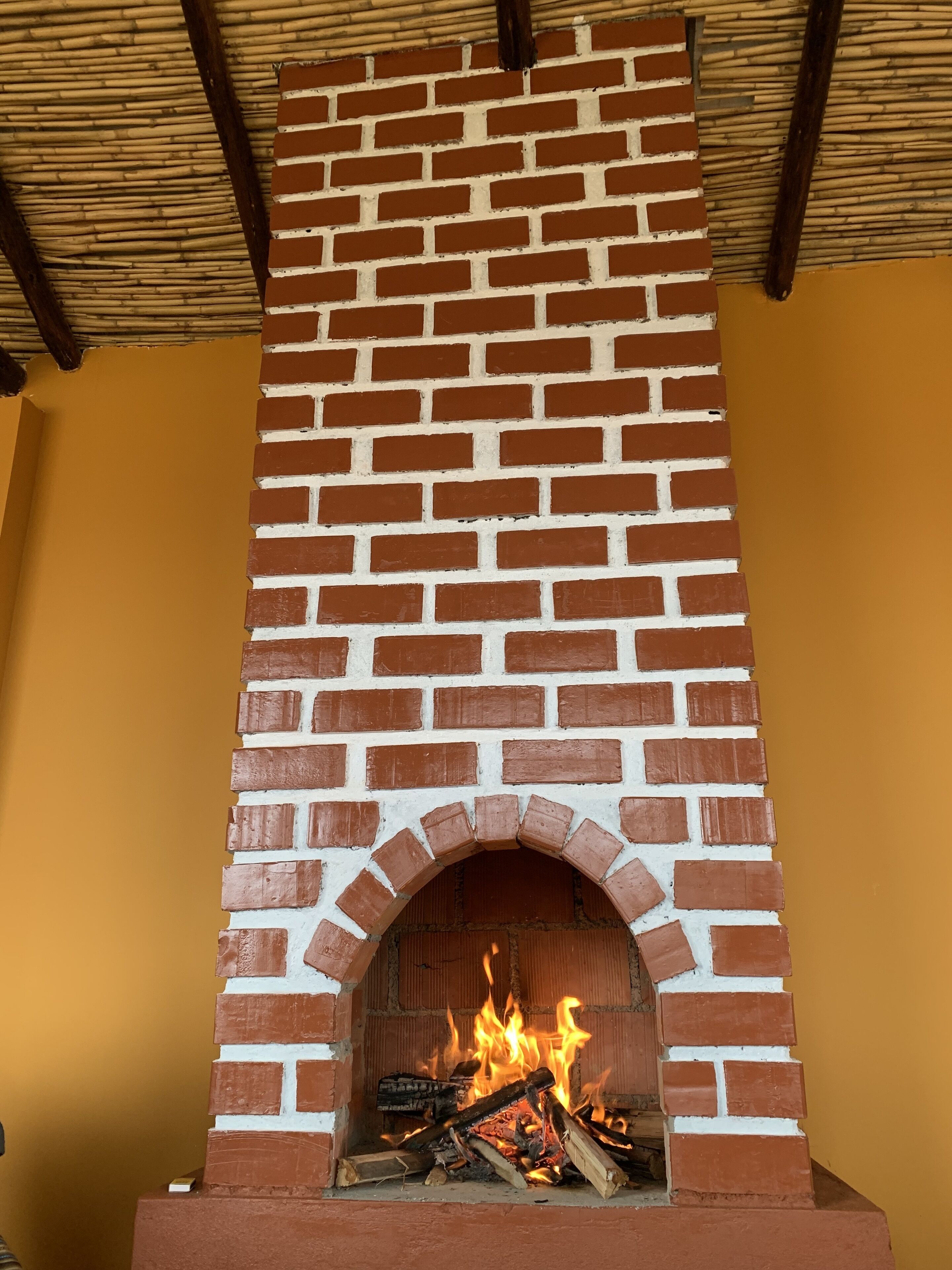 A brick fireplace with a fire burning inside, set in a room with yellow walls and a wooden ceiling.