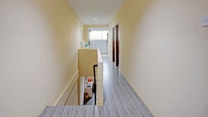 A narrow hallway with a staircase on the left, leading to a landing with a small table and a chair. The walls are painted in a light color, and the floor has a patterned tile design. - Hotel O Aulia Homestay Syariah (Batam)