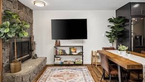 A cozy living room with a fireplace, a flat-screen TV mounted on the wall, a bookshelf, and a dining area with a wooden table and chairs.